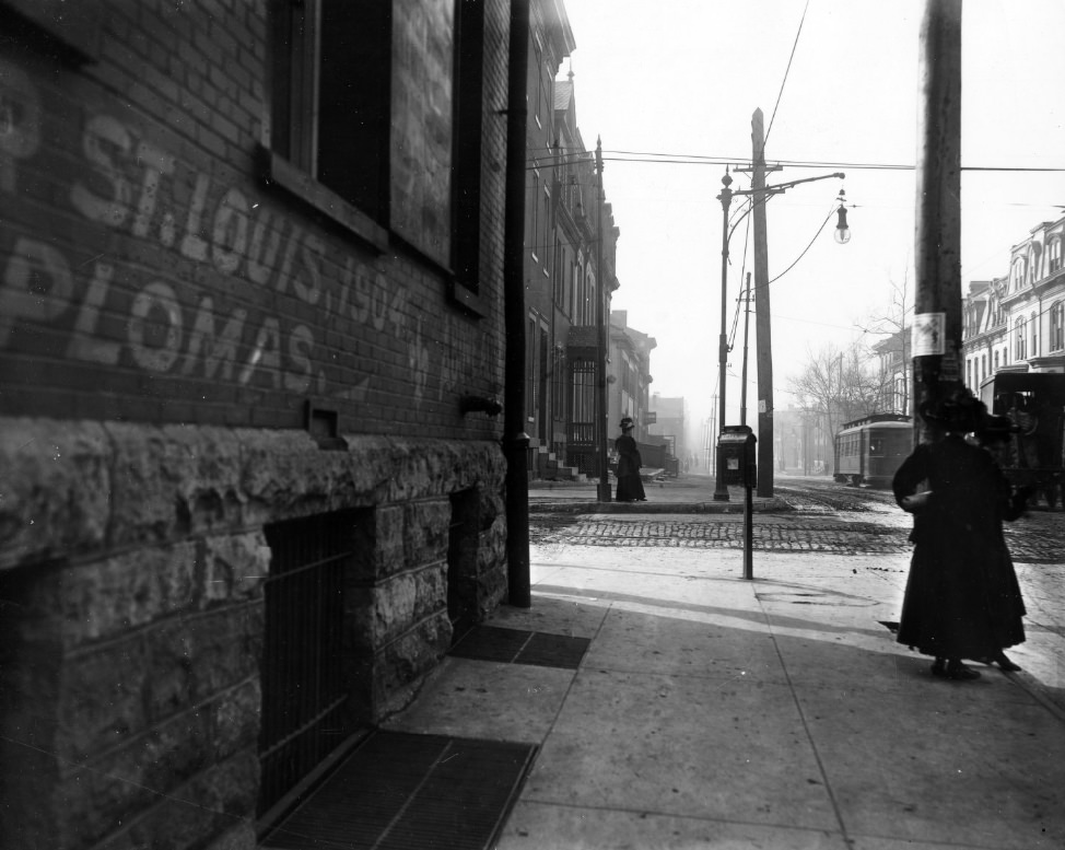 #92 Street scene of St. Louis after during or after 1904. Two women on sidewalk. Trolley car on rails in the cobblestone street. Buildings with mansard roofs.