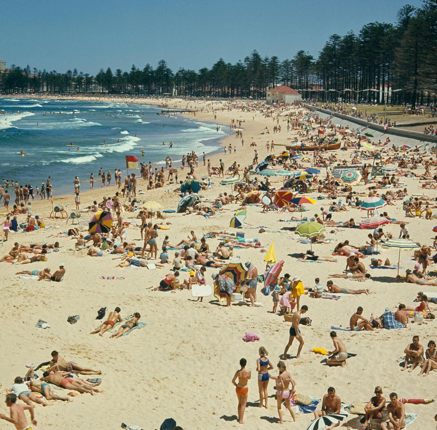 #87 Visitors sunbathe under umbrellas on the sandy beach facing the Pacific Ocean in the suburb of Manly in northern Sydney, 1965