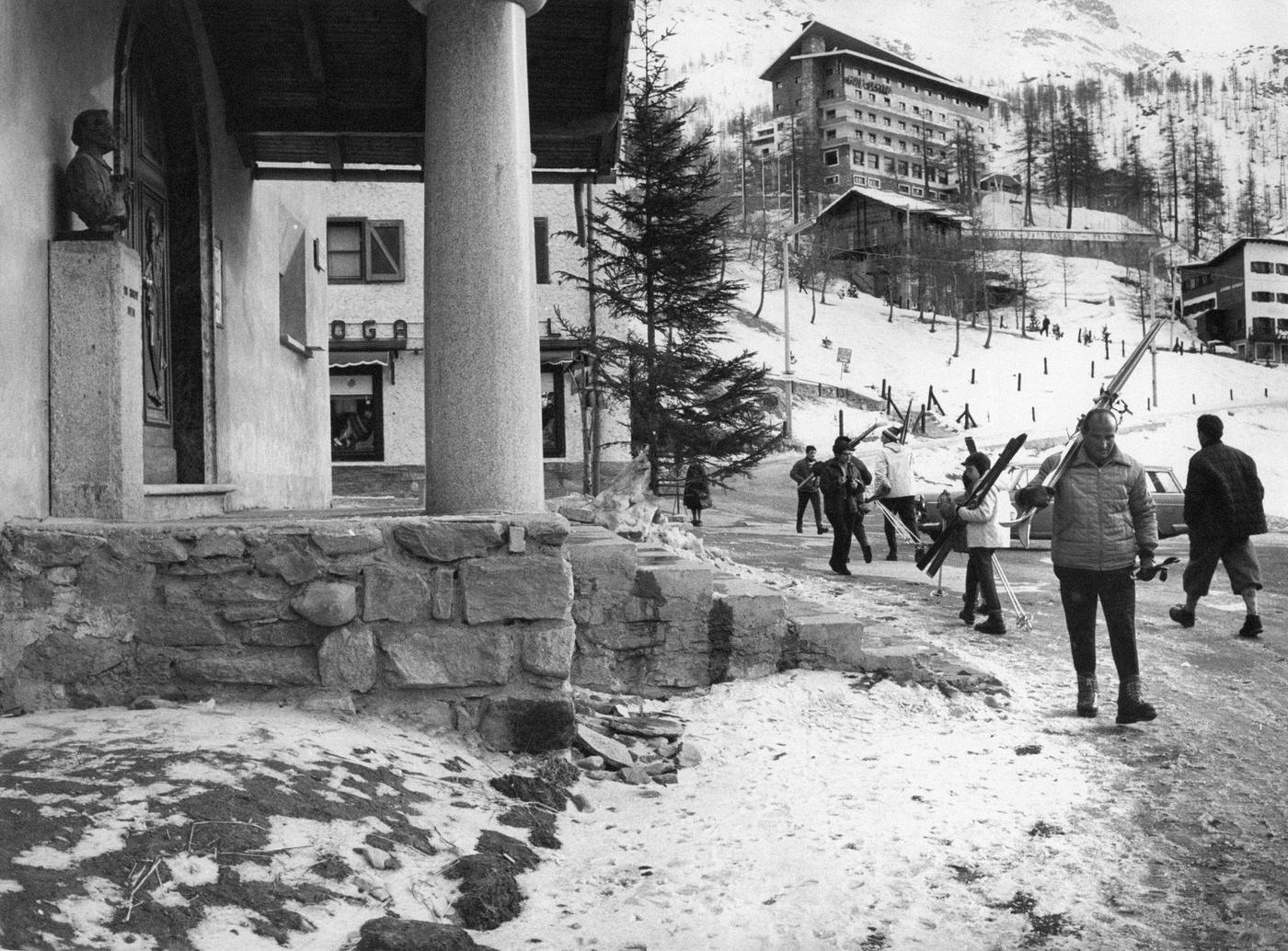 #35 Some skiers, with the skis on their shoulders. walking in front of the new church founded by Don Giuseppe Vietto, the odd priest of Cervino. Valtournenche, 1964