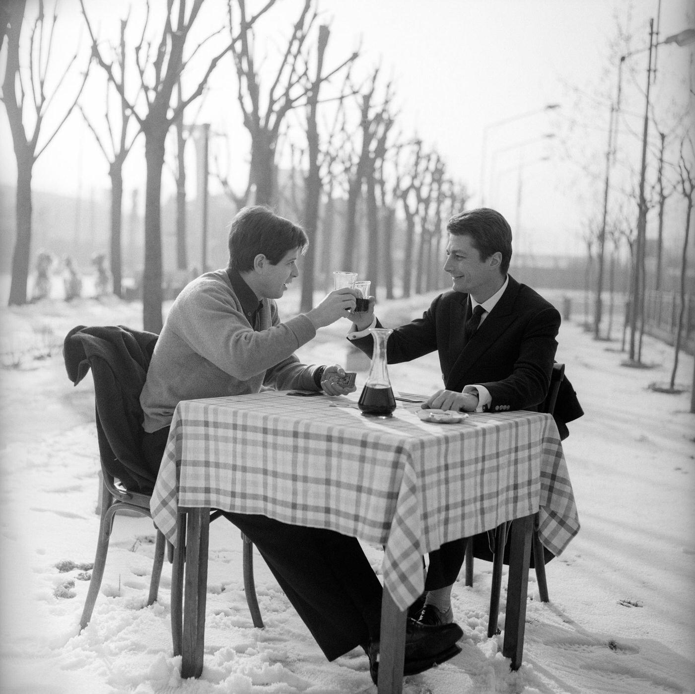 #38 The Italian voice artist and film actor Corrado Pani and the US actor Joseph Walsh are chatting around a square table in the open air, in a snow covered landscape; they are toasting with red wine. Italy, 1963.