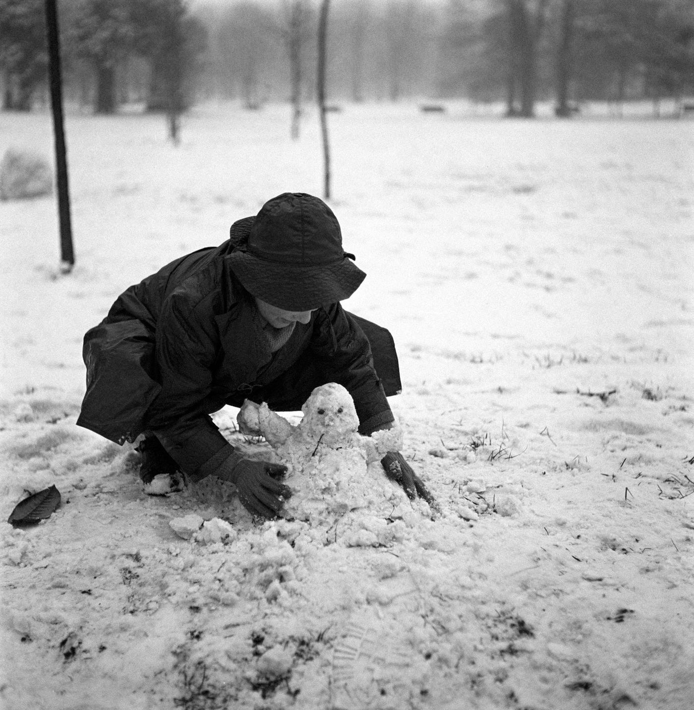#53 Little boy crouching and making a snowman in a park after a snowfall. Milan, 1950s.