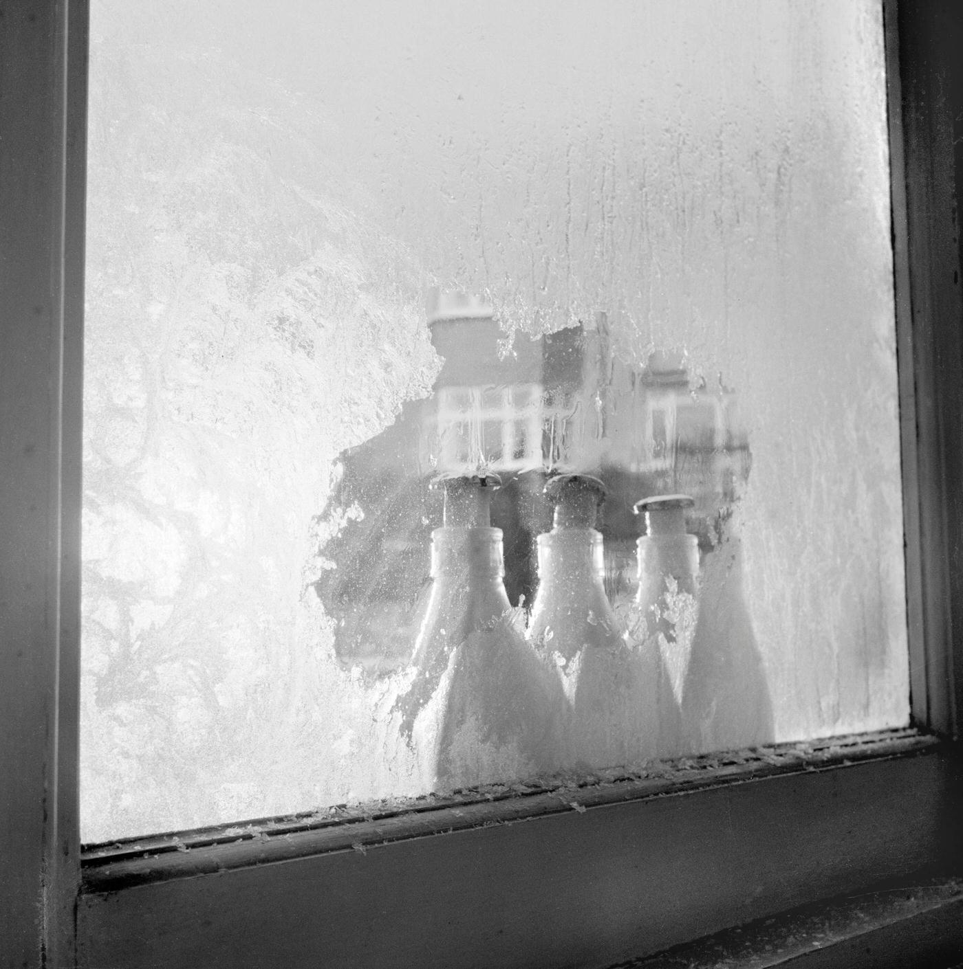 #13 A row of three milk bottles standing outside a window with frozen milk pushing the caps off, 1960s