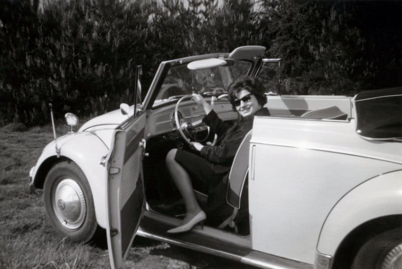 A cheerful lady posing in the driver’s seat of a VW 1200 Cabriolet in the countryside. The car is equipped with fender guides and an additional wing mirror, 1963