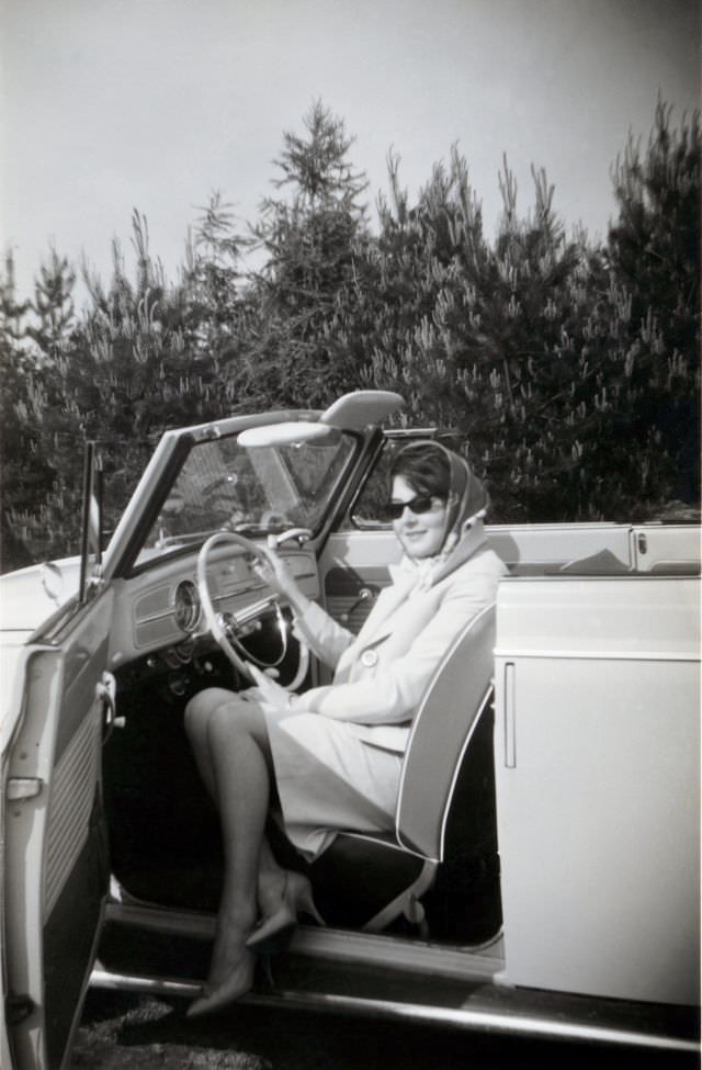 An elegant lady posing in the driver’s seat of a VW 1200 Cabriolet in the countryside. The print is dated “1963” on reverse