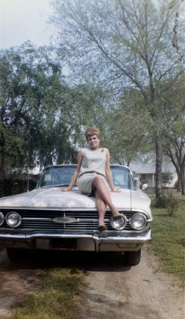 A young blonde lady in a sleeveless white dress posing on the bonnet of a 1960 Chevrolet Impala in the drive of a private home. The words “Nancy 1964” are handwritten on reverse