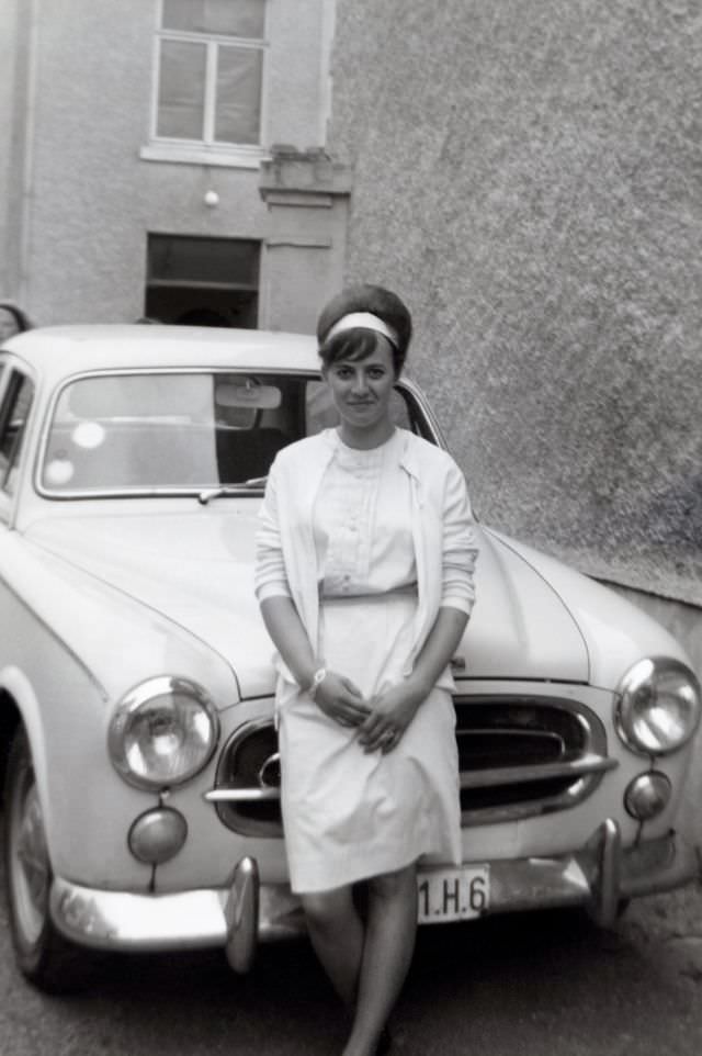 #42 A cheerful lady with a beehive hairdo posing with a Peugeot 403 in an urban backyard. She is wearing a white skirt, blouse, cardigan and hairband. The car is registered with Belgian licence plates, 1960s