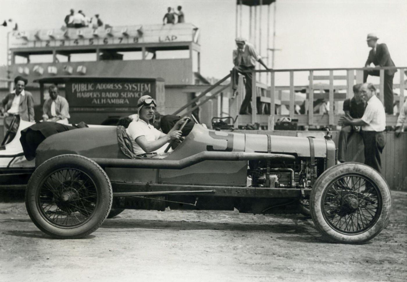 #128 Joe Crocker poses in a race car at the speedway with a group of spectators milling about in the background, 1935.