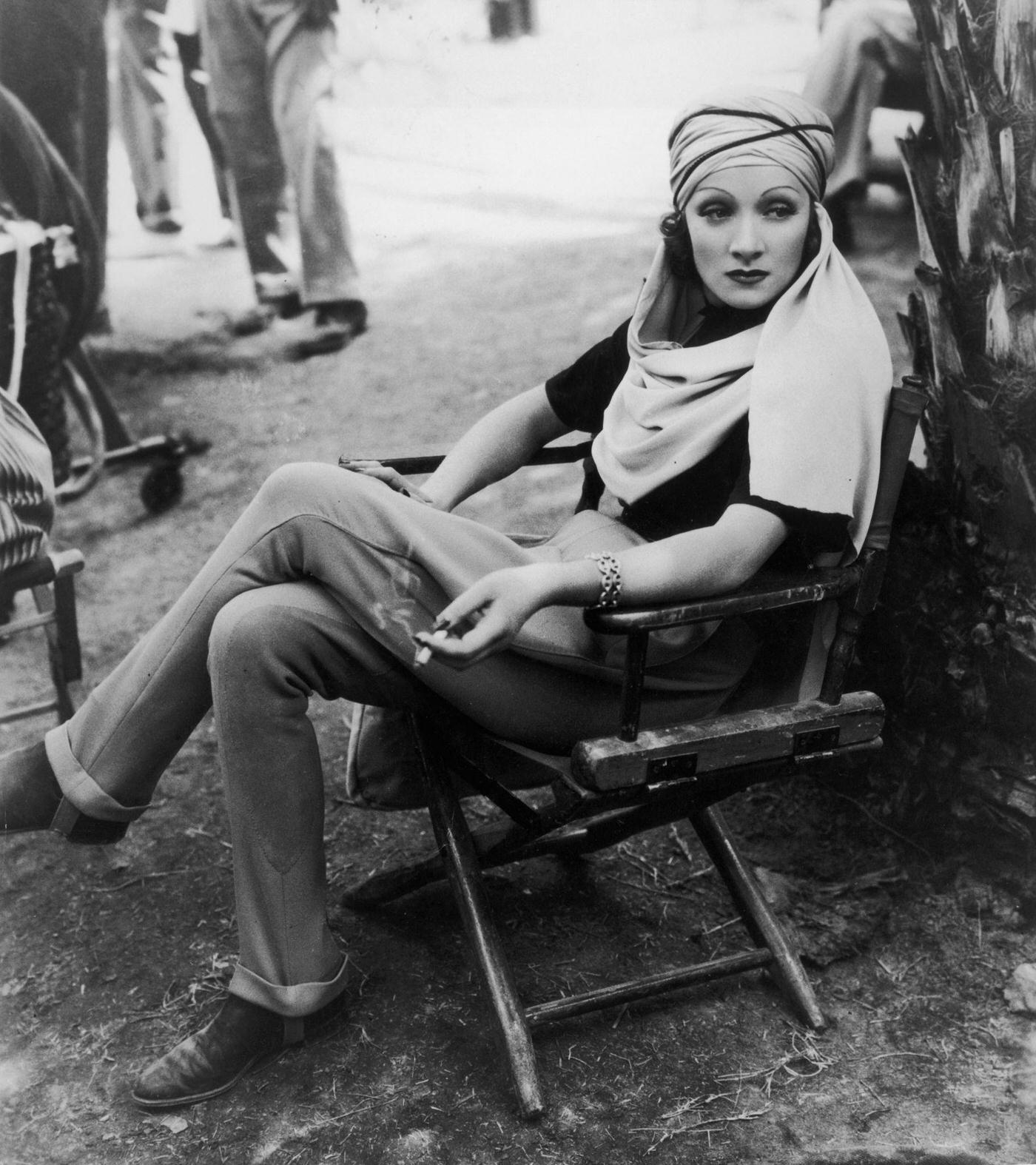 #10 Marlene Dietrich sits in a canvas chair, wearing a turban, and holding a cigarette on the desert location set in 1936.