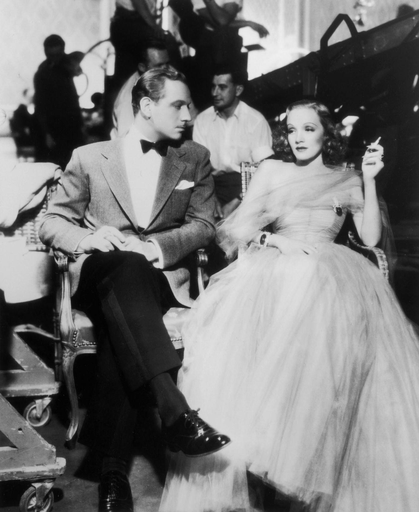 #19 Melvyn Douglas sits next to German-born actress Marlene Dietrich as she smokes a cigarette in 1937.