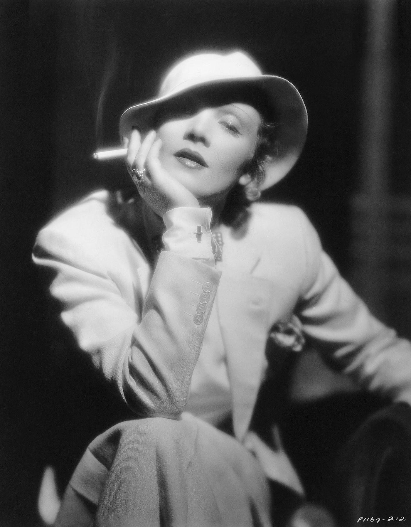 #33 A photograph of a German-born Marlene Dietrich holding a cigarette, dressed in a tailored suit, hat, bow-tie, and cufflinks.