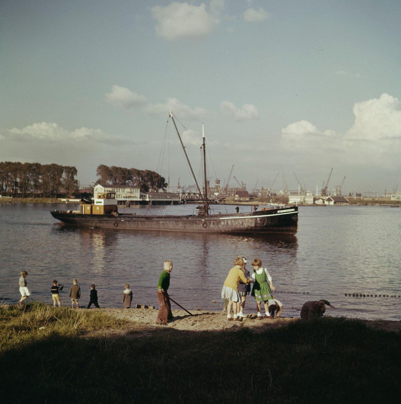 #46 A barge passes children playing on a canal bank in a suburb of Amsterdam, capital city of the Netherlands, 1950.