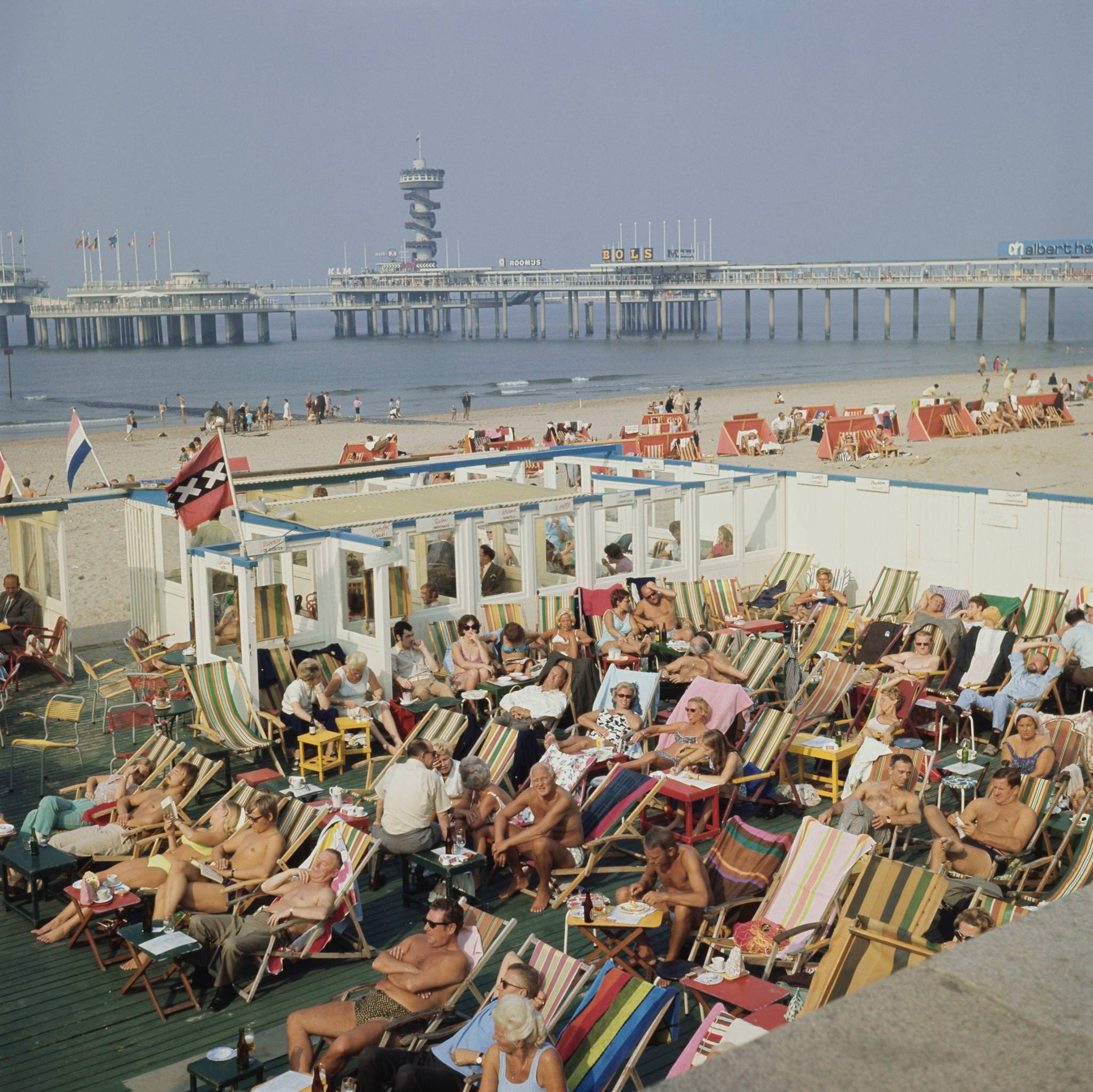 #74 Visitors and holidaymakers sunbathe on deckchairs on a terrace beside the sandy beach at the seaside resort of Scheveningen near The Hague in the Netherlands, 1970s