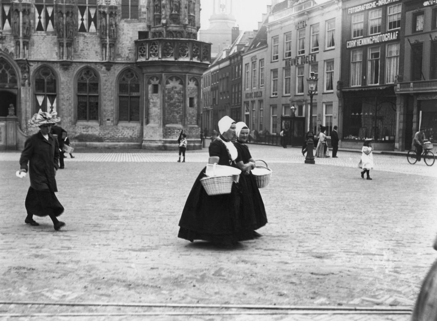 #85 Pair of Dutch women carrying baskets crossing a town square, Netherlands, 1900