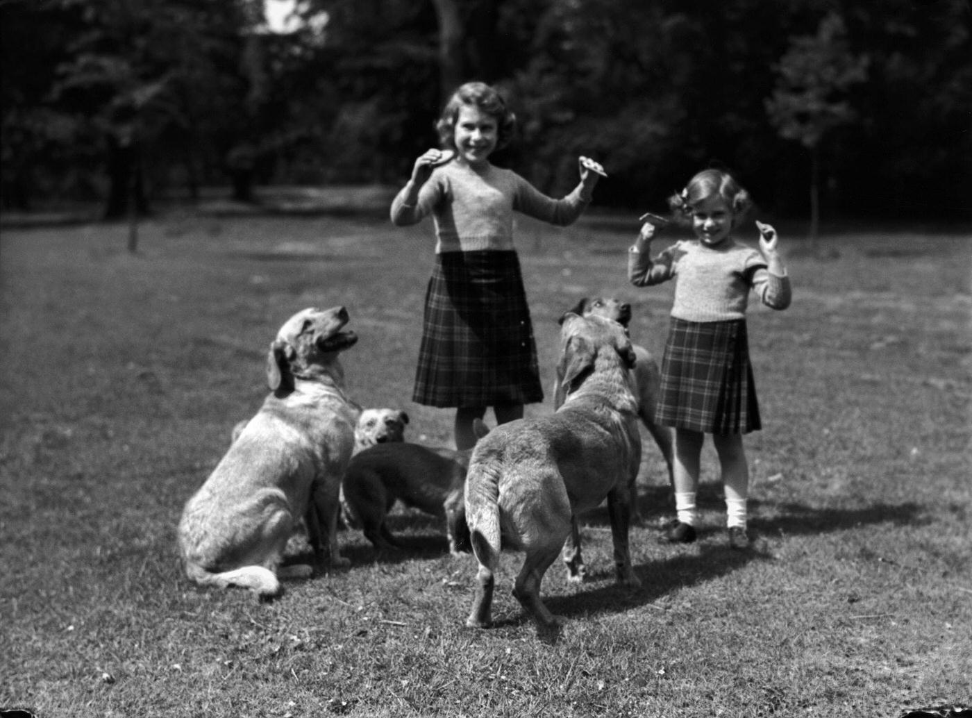 #101 Princess Elizabeth and her sister Princess Margaret wearing kilts and feeding biscuits to dogs at the Royal Lodge, Windsor, UK.