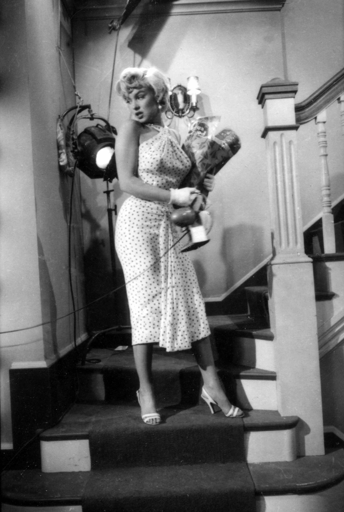 Marilyn Monroe, wearing a polka-dot dress, ascends a staircase holding a bag of groceries and a desk fan in her arms in 1954 during the filming of “The Seven Year Itch”