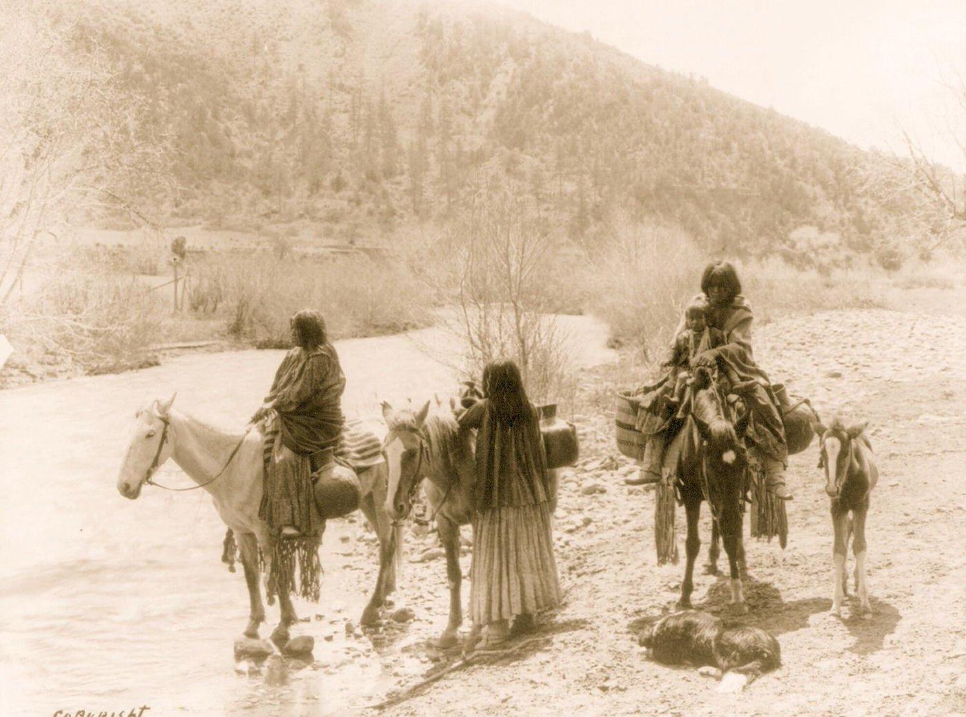 #20 Three Apaches, one with a child, with horses laden with water jugs, a colt, and a dog stopped beside a stream, 1903