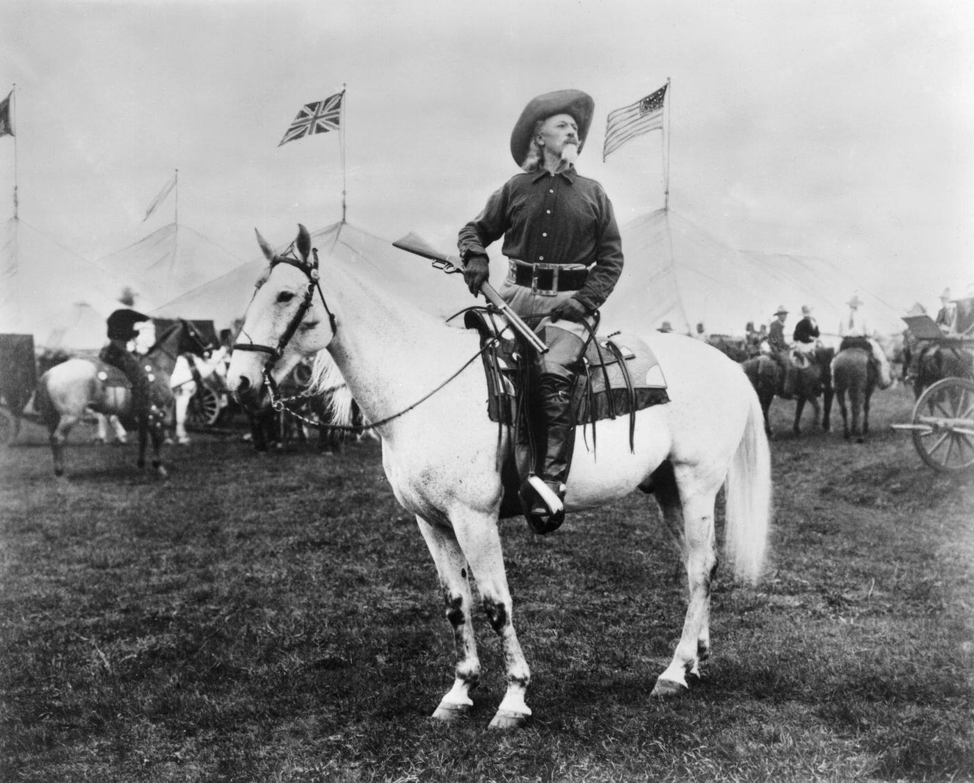 #29 William ‘Buffalo Bill’ Cody (1846 – 1917) American entertainer, sitting on horseback and holding a rifle, looks off into the distance as British and American flags fly around him. Tents for his Wild West show are in the background.