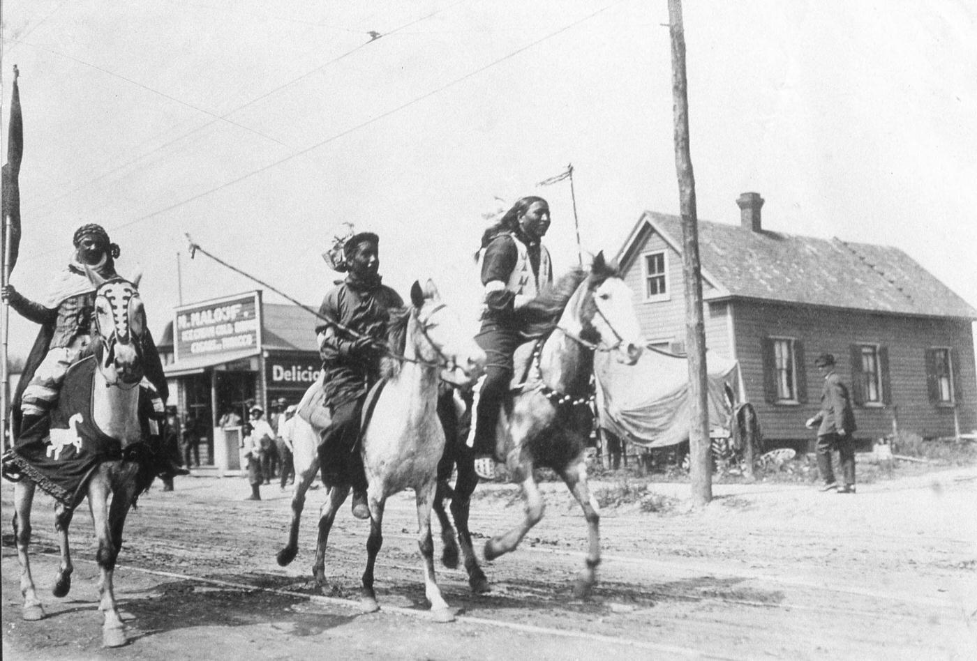 #32 A photograph shows three Native American men riding horses during Buffalo Bill’s Wild West Show, United States, late 19th century. They race down a road in a rural town.