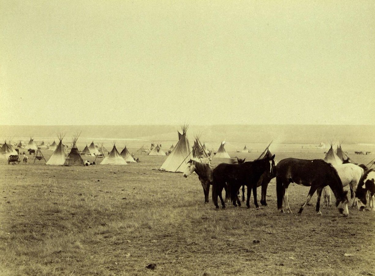 #59 Blackfoot Camp, Blackfeet Indian Reservation, Montana, 1887: A group of horses graze in the foreground as a Blackfoot Indian encampment spreads into the background behind them. Text reads ‘Indian Camp, Blackfoot Reserve.’