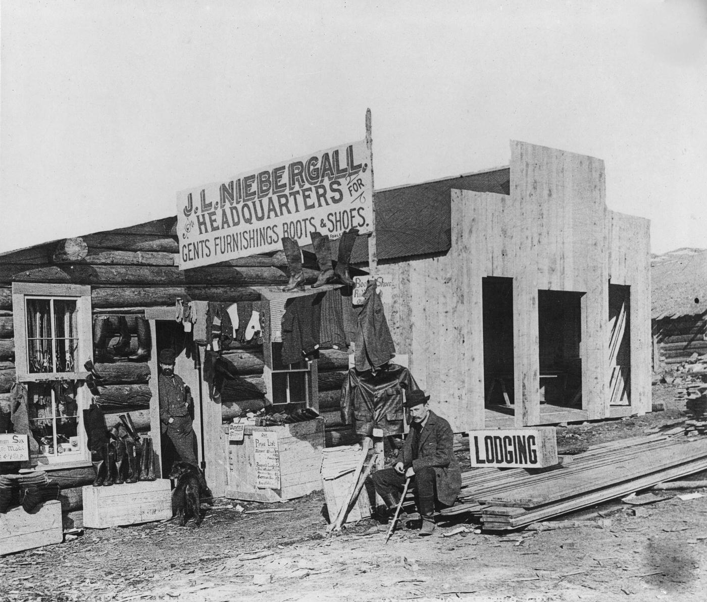 #72 J.L. Niebergall, Headquarters For Gents Furnishings Boots & Shoes, exterior view, with two men visible, of JL Niebergall, billed as the headquarters for Gents Furnishings Boots & Shoes.