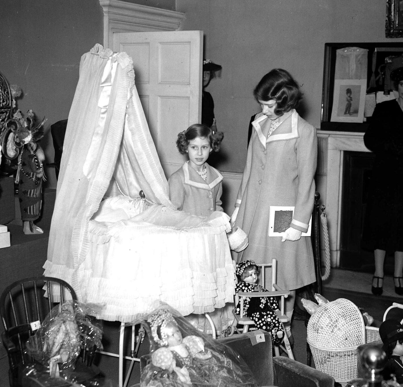 #17 Princesses Elizabeth and Margaret view the cot that they both used as babies during their visit to an exhibition of royal and historic treasures at their old home, 145 Piccadilly, London, 28th July 1939.
