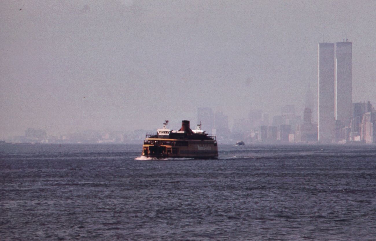 #103 Staten island ferry with smog-obscured skyline of lower manhattan in background. On the right are the twin towers of the world trade center overlooking the hudson river, 1970s