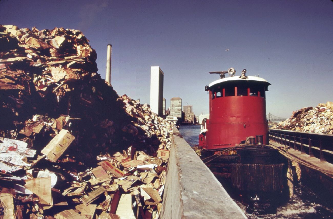 #87 Tugboat herds two laden garbage scows down the east river from transfer point at 91st street. Destination is the staten island landfill, 1970s