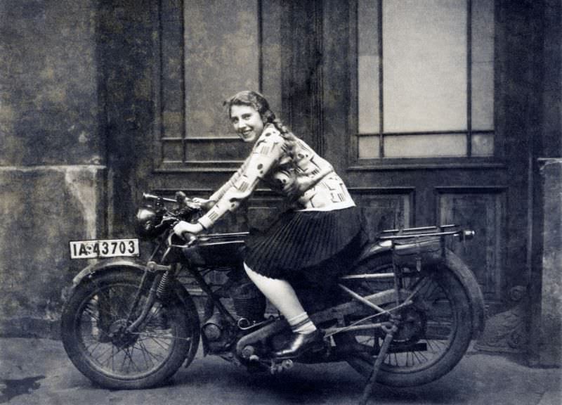A cheerful young lady posing in the saddle of an ancient motorcycle in front of a double-leaf door. The words “1 january 1930” are handwritten on reverse. The bike is registered in the city of berlin.