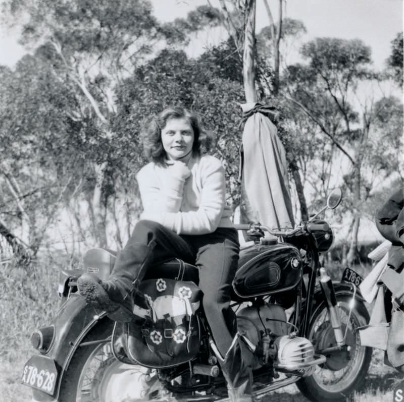 A young lady wearing a white turtle-neck sweater, stirrup pants, and suede leather boots posing in the saddle of a bmw r50/2, circa 1965.