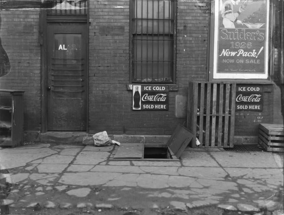 #64 21st and Chestnut, 1930 – View of an open cellar door behind a grocery store. Advertising for Coca-Cola and Snider’s ketchup are hung on the building.
