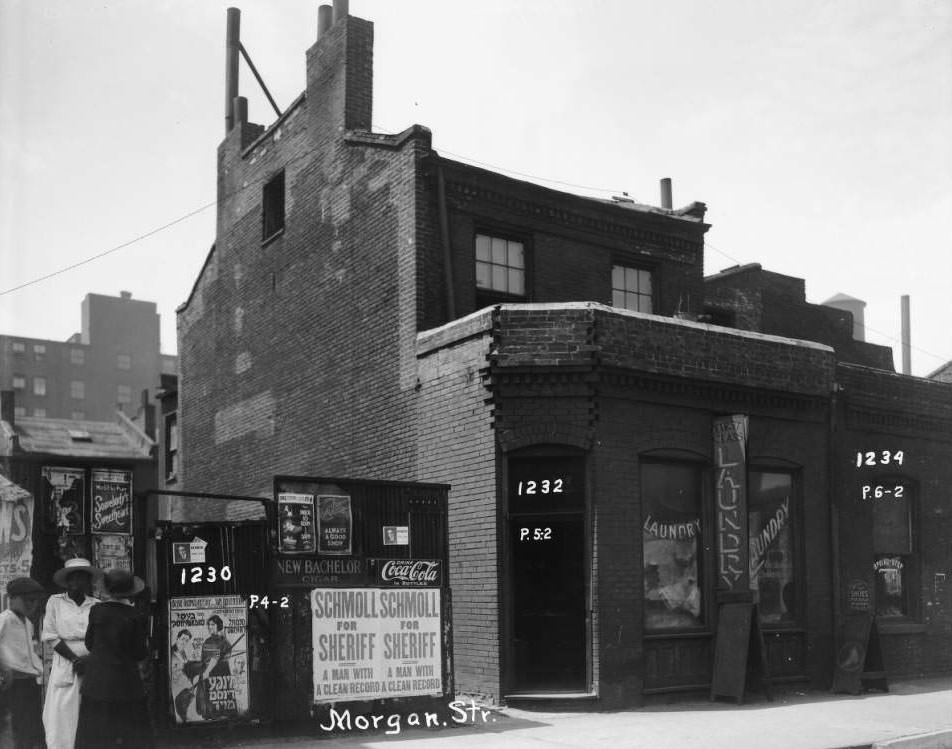 #78 Exterior view of a laundromat and shoe repair shop with three people standing in front of billboards. 1232 and 1234 Morgan St, 1930