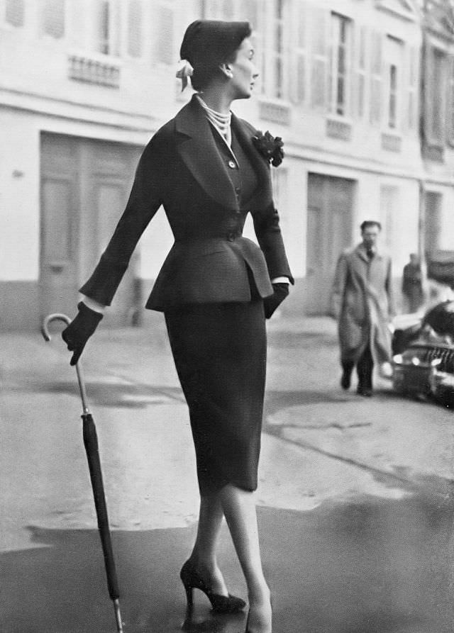 #8 Barbara Goalen in fitted navy blue wool suit with horseshoe neckline filled in with matching vest, skirt is made up of two petal-shaped pieces, by Christian Dior, 1950.