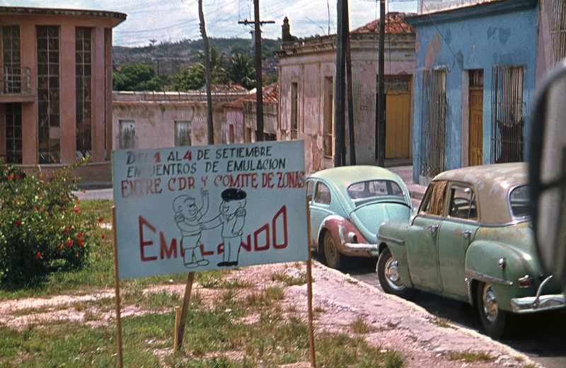 #39 Old American cars parked on a street with old houses. In a sign at the street: “Emulation Meetings between CDR and Zoning Committee announced”, Cuba, 1976