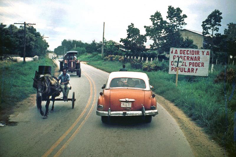 #40 On the road, an old American car, a horse and cart and a tractor. Sign near the road of socialist government message: “To decide and govern with the people’s power”, Cuba, 1976