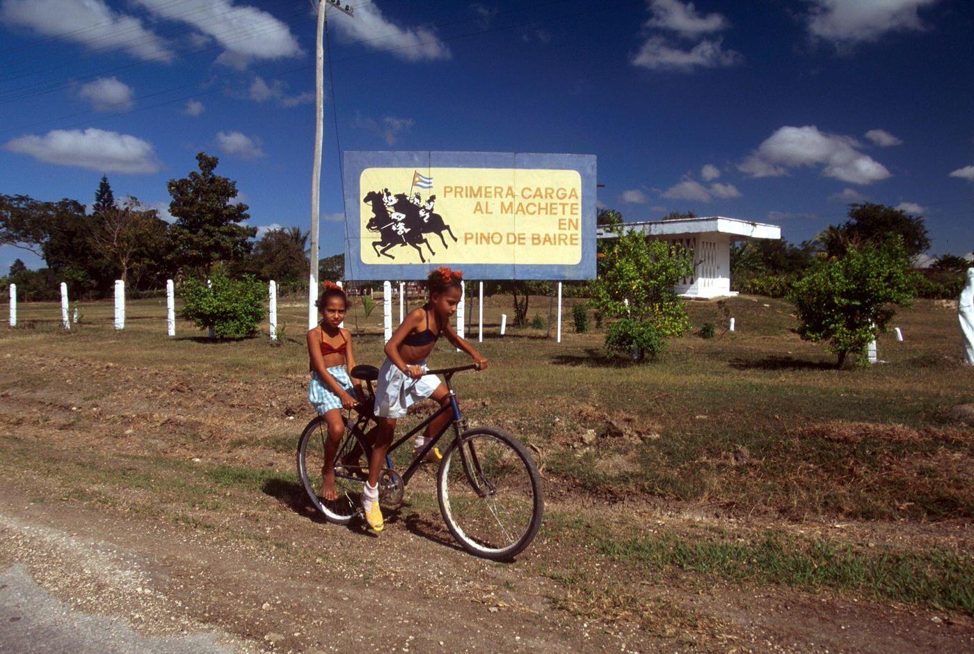 #34 Two Cuban girls riding bicycles with a poster commemorating a historical event of the Cuban War of Independence, Cuba, 1990.