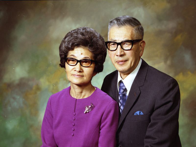#19 Mr and Mrs Kinoshita. Mrs Kinoshita is wearing a purple top with a metal pendant. Mr. Kinoshita is wearing a white shirt, blue-silver tie, and a charcoal suit jacket. Both individuals are wearing black-framed glasses, February 1973