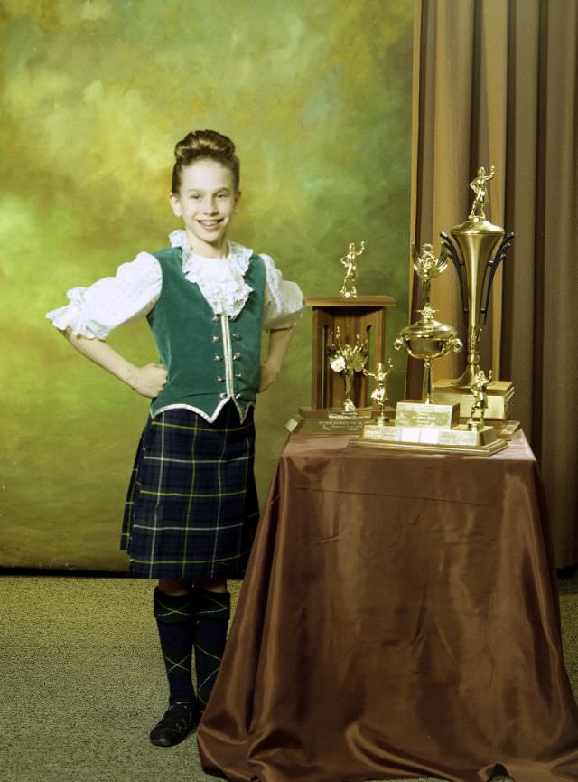 #30 Portrait of a girl posing with three trophies. The girl is dressed in a Highland dance outfit and the trophies have been placed on a low, draped table on the right-hand side of the image, March 29, 1978
