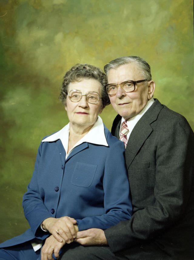 #33 Portrait of an older couple. The woman is wearing a blue pant suit over a white shirt with a broad collar. The man is wearing a charcoal grey suit over a white shirt and plaid tie. Both individuals are wearing glasses, November 22, 1978