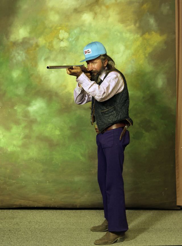 #35 Portrait of a man in purple pants, a turquoise cap, and a long-sleeved light shirt. He is holding a gun, and standing before a green background, 1979