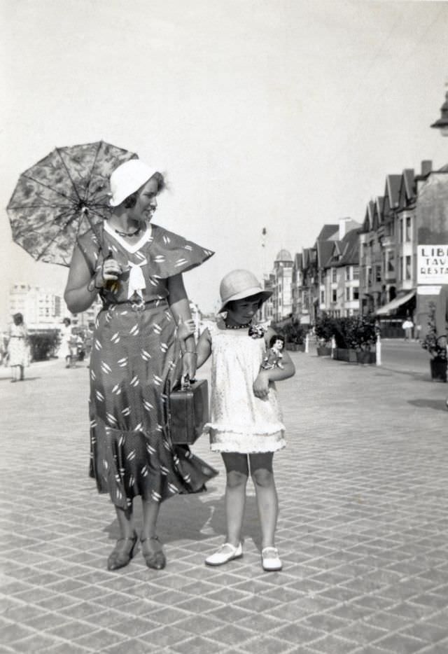 #5 A fashionable lady carrying a lace parasol and her daughter, holding a doll in her arm, posing on a seaside boulevard in summertime, 1930s