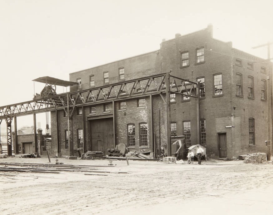 #113 Small factory or warehouse on First Street with an apparatus for loading materials onto rail cars attached to the front of the building and two workmen relaxing in front of it, 1920