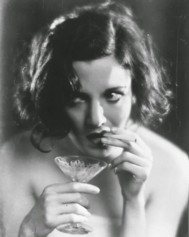 #7 A woman smokes a cigarette while holding a cocktail glass in the other hand, 1929