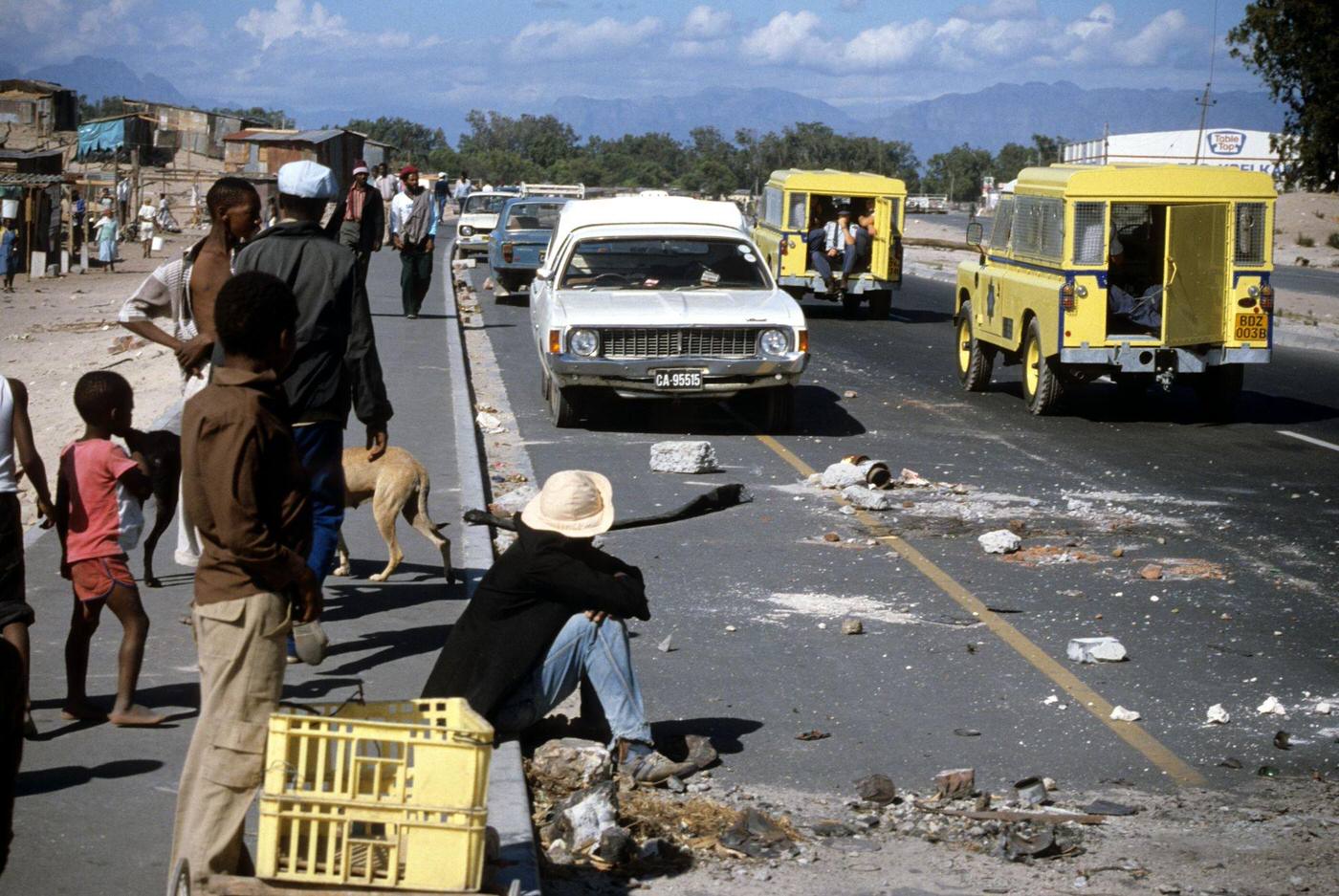 #80 People on a sidewalk watch as South Africa Police Land Rovers drive past debris on a road in the Crossroads shantytown, Cape Town, 1985