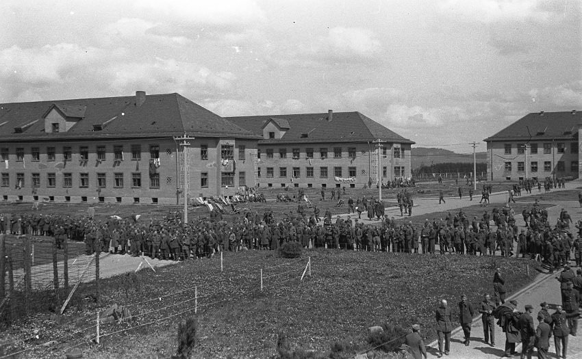 #19 A relaxed atmosphere pervaded in the camp after its liberation by American forces in 1945. In front of the barracks on the left, some former inmates sit on lounge chairs in the sun.