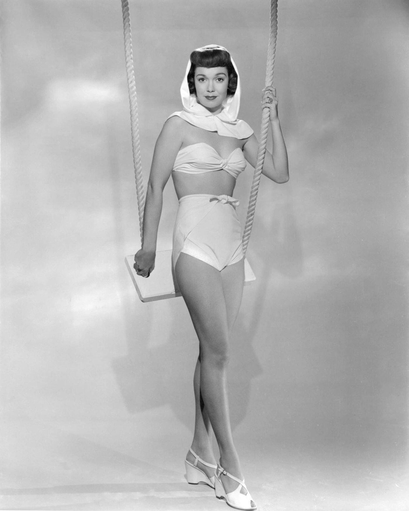 #32 Jane Wyman, American actress and singer, leaning on a swing in a white two-piece swimsuit, 1945.
