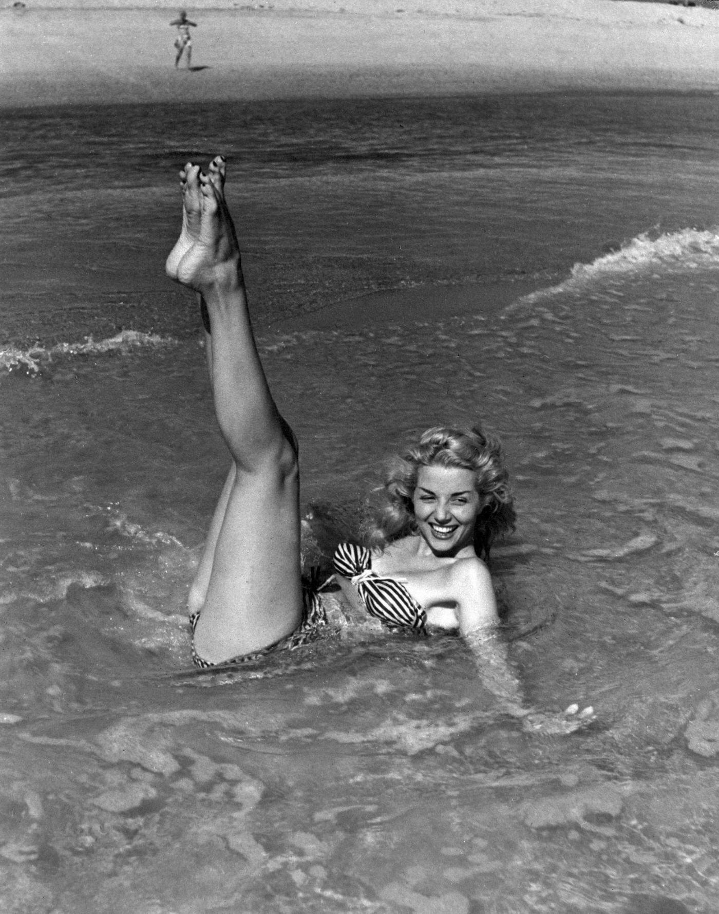 #56 Young woman laying on the seashore wearing a striped bikini, 1948.