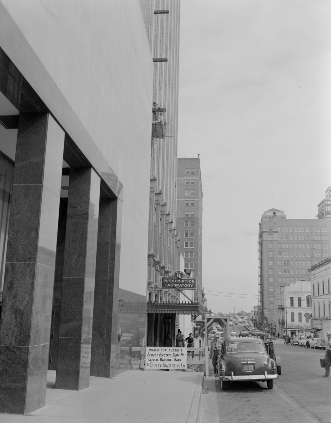 #48 Busy Street Flanked by Large Buildings Including a Pharmacy, 1950s.