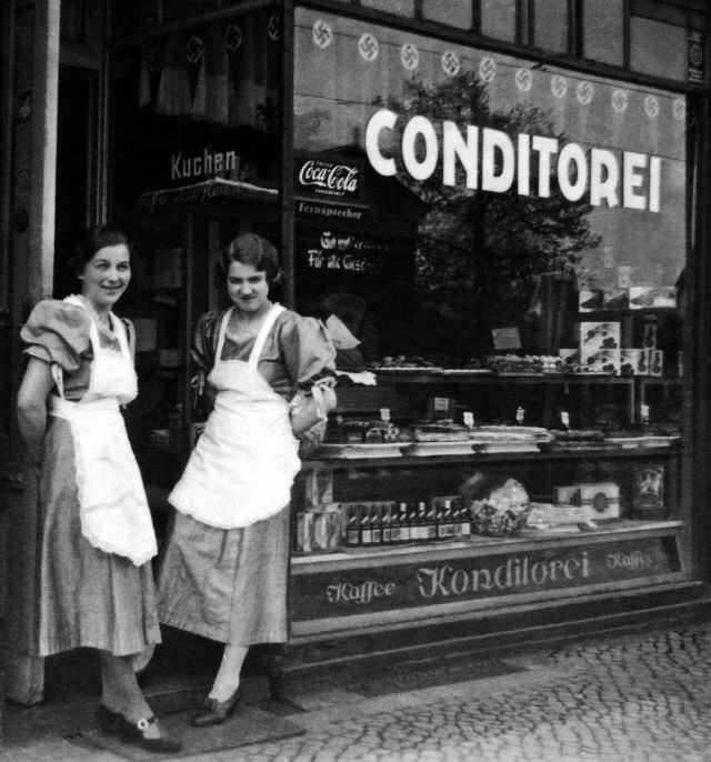 #39 Two ladies pose in front of a Konditorei in Germany displaying a Coca-Cola sign, circa WWII