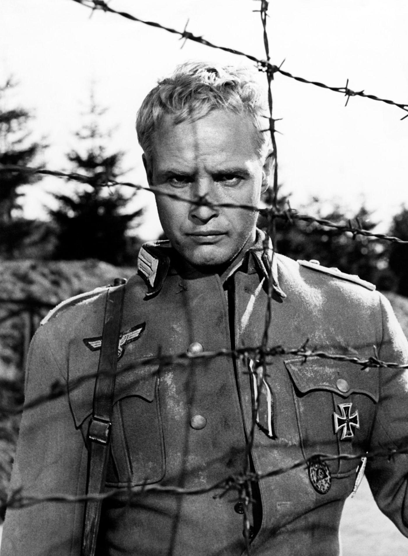 #58 Marlon Brando behind barbed wire, shooting “The Young Lions,” Natzweiler-Struthof Concentration Camp, France, 1957