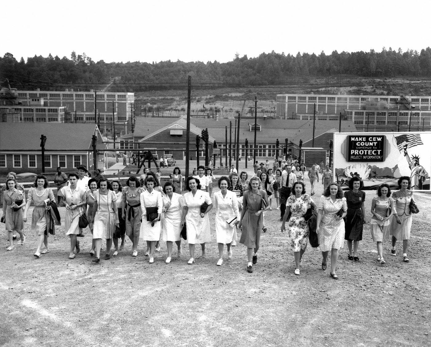 #23 Oak Ridge Shift Change at Y-12 Uranium Enrichment Facility. 1944. Billboard reads “Make CEW count Continue to protect project information.